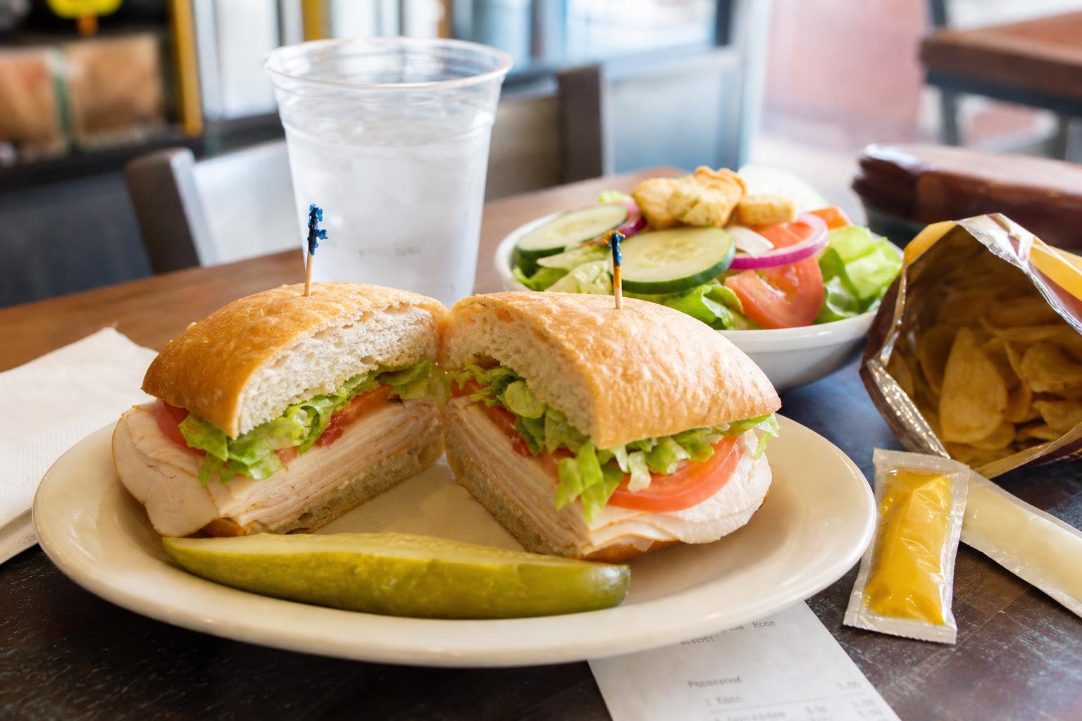 Realistic sandwich shop lunch with a cut deli sandwich, side salad, chips, pickles, condiments, and water in warm daylight