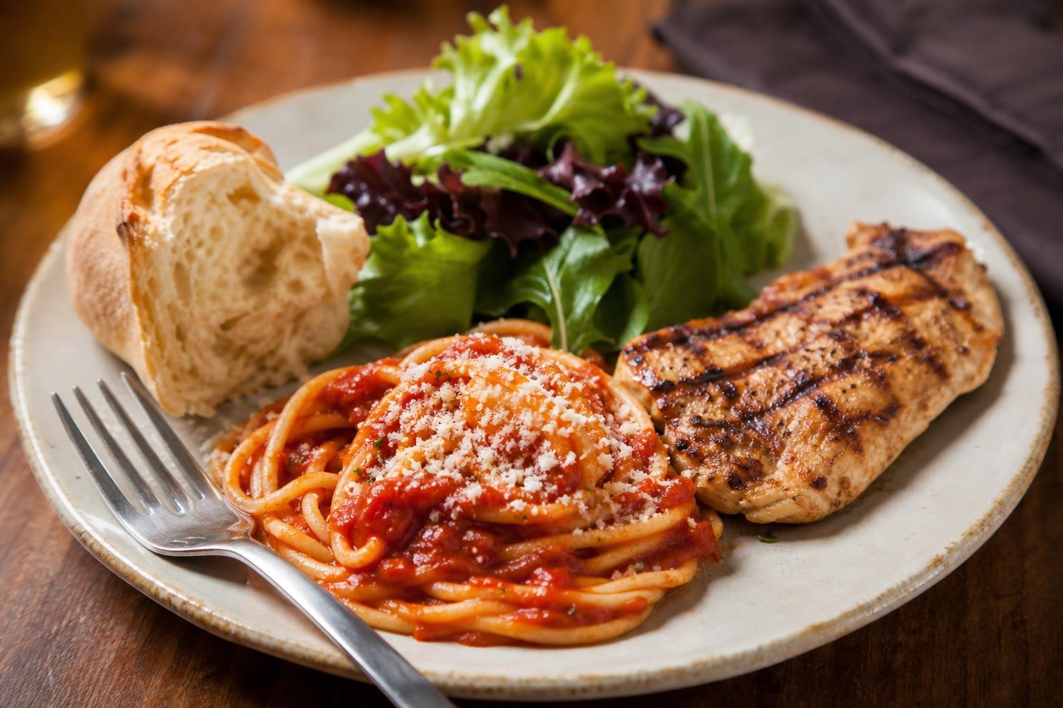 Close-up photorealistic plate of pasta with grilled chicken, parmesan, and a smaller bread portion on a restaurant table