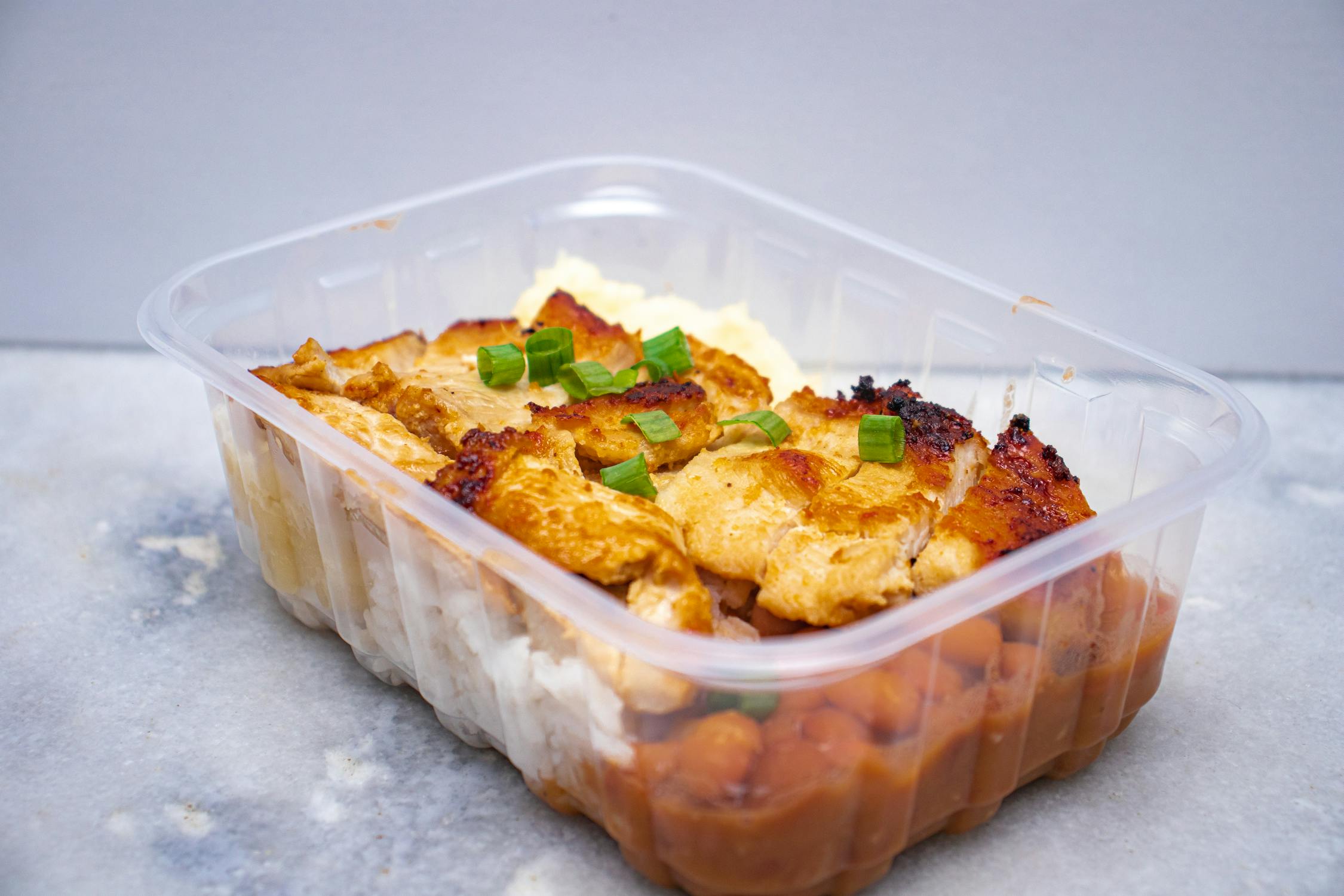 Grilled chicken rice and beans packed in a clear takeout container on a dark table